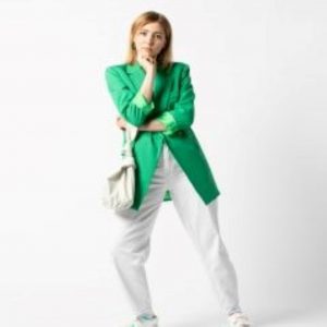 Model wearing a green coat and white trousers against a white photographic backdrop